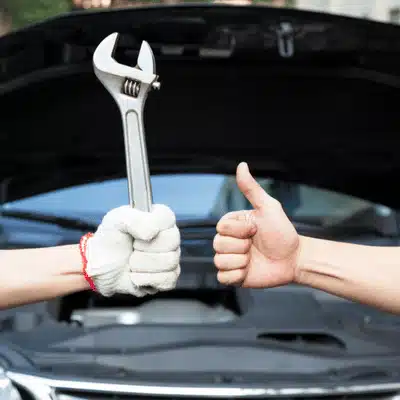 Gloved hand holds wrench while another gives thumbs-up before car with open hood after repairs
