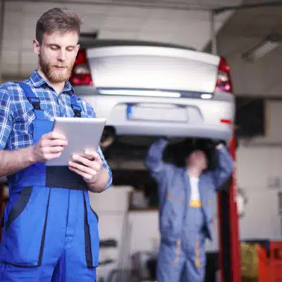 Auto mechanic reviewing service data on a tablet while another mechanic works under a raised vehicle
