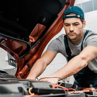 Auto technician inspects engine components with hood open, performing diagnostics and testing electrical connections carefully
