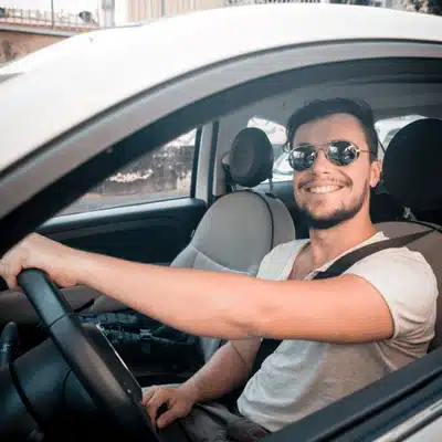 Driver wearing sunglasses smiles from car seat, buckled and ready for safe, comfortable summertime journey